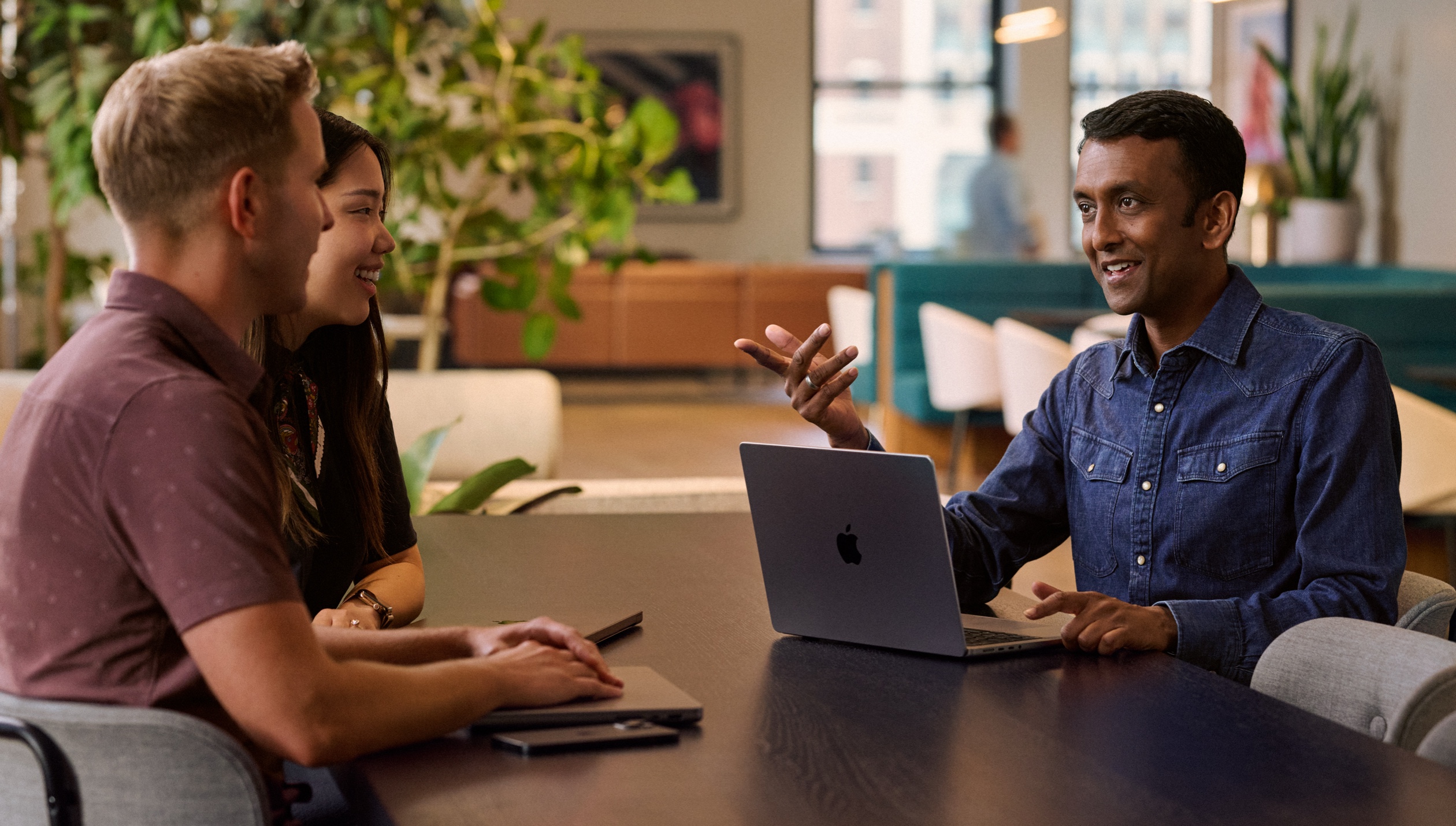 Three colleagues in a discussion sit at a table with their MacBooks and an iPhone in front of them.