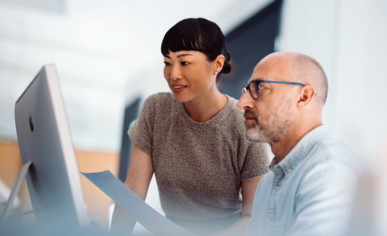 Two colleagues, one standing and one seated, look at the display of an iMac.