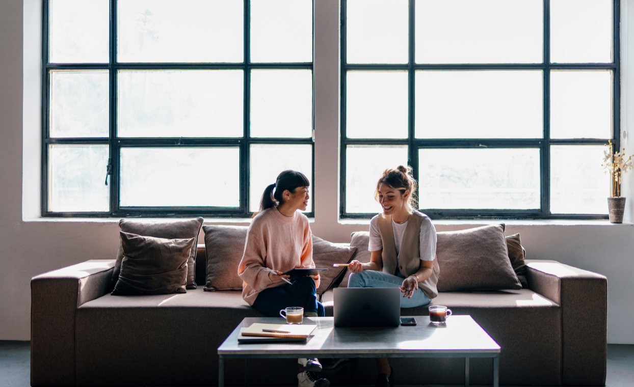 Two colleagues seated on a couch with a MacBook and two drinks in mugs in front of them.
