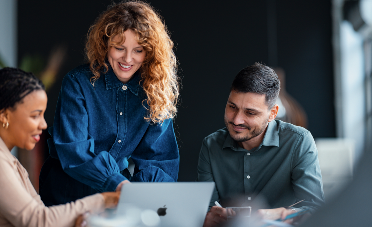 Three colleagues, two seated and one standing, look at the screen of a MacBook.