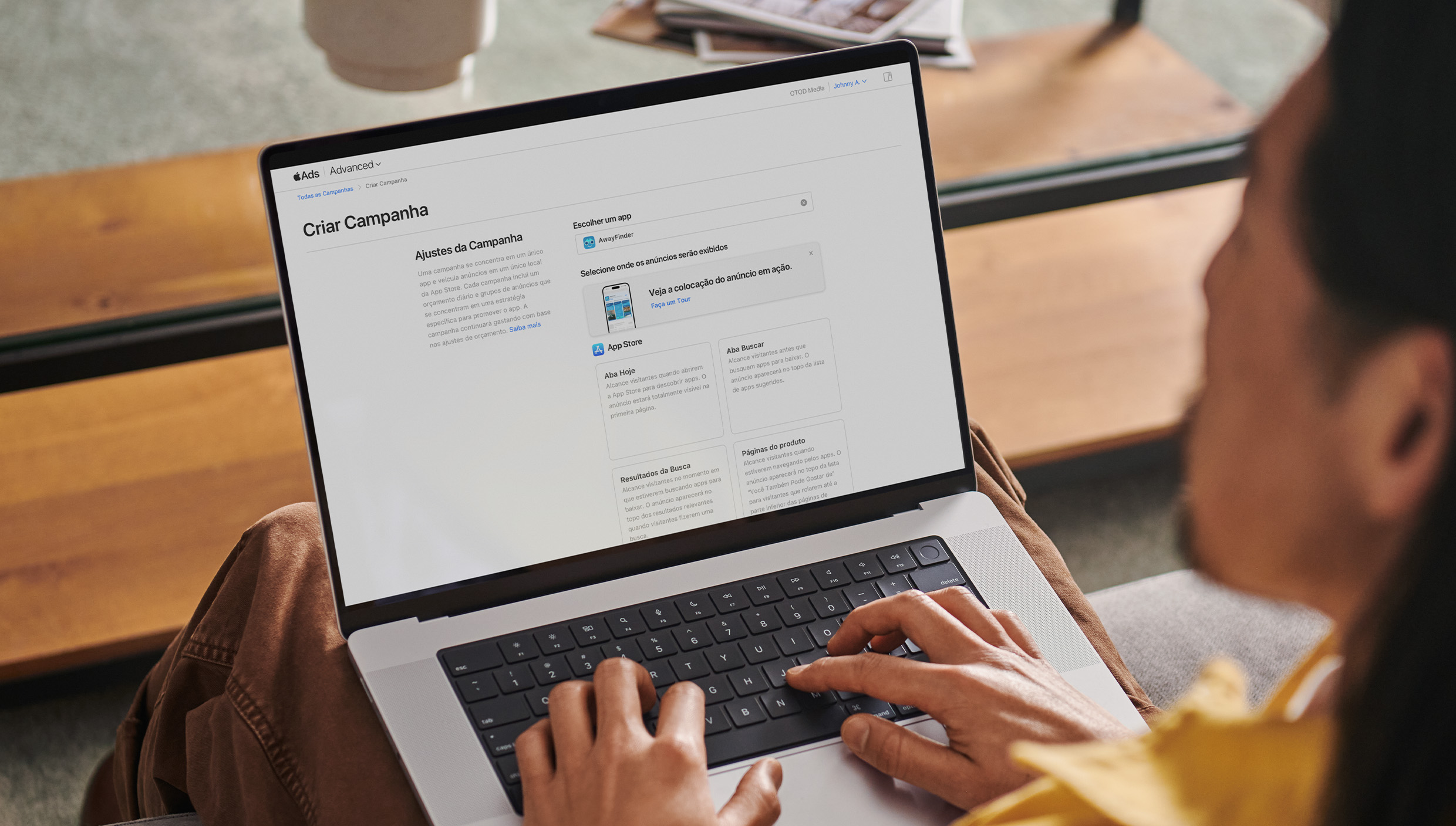 Anunciante trabalhando em um MacBook. A página Criar Campanha do Apple Ads está aberta na tela exibindo quatro colocações de anúncios disponíveis: aba Hoje, aba Buscar, resultados de busca e páginas de produtos.