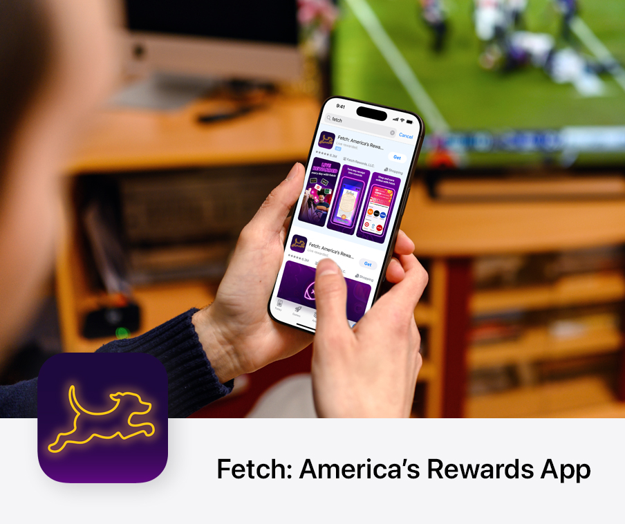 A person holds their iPhone while they sit in front of a TV that shows a football game being played. On their iPhone, a search results ad for the Fetch app appears on the App Store.