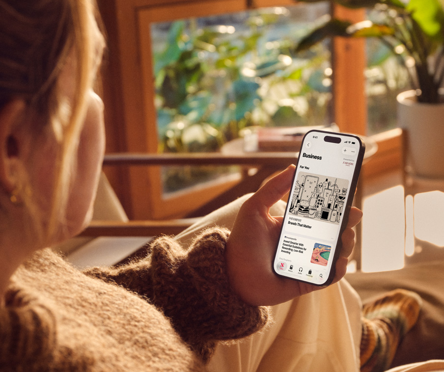An over-the-shoulder image of a person sitting on a couch looking at an Apple News article on their iPhone.