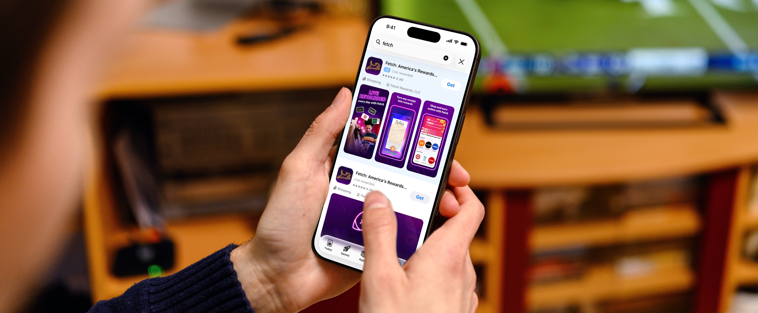 A person holds their iPhone while they sit in front of a TV that shows a football game being played. On their iPhone, a search results ad for the Fetch app appears on the App Store.
