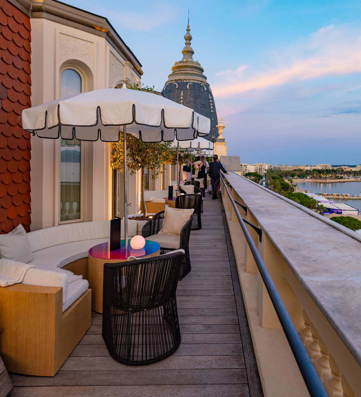 A sunset view of the balcony at the Carlton in Cannes, with umbrella tables set up and the beach and ocean below.