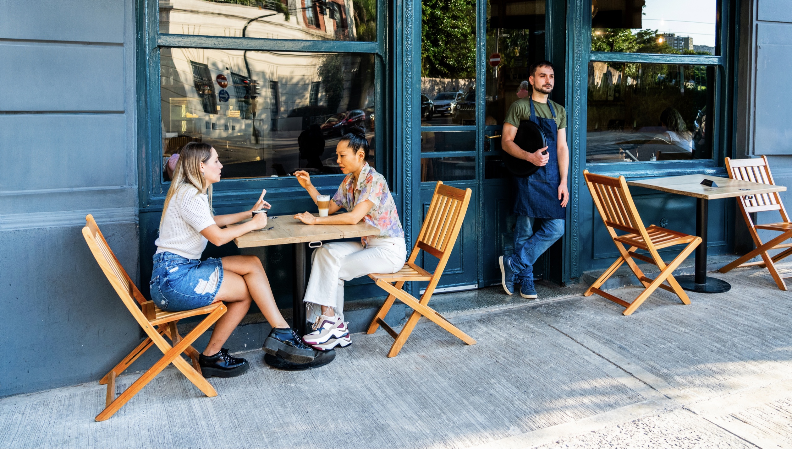 Two people sit and enjoy coffee outside a shop. Employee looks out in the doorway.