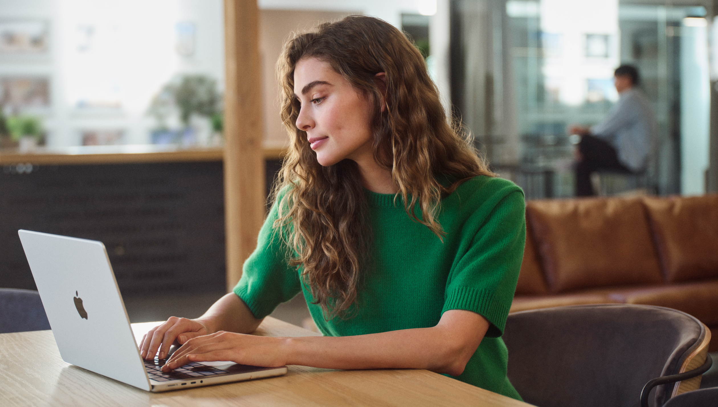 A person types on a MacBook at a wooden table in a modern, open workspace.