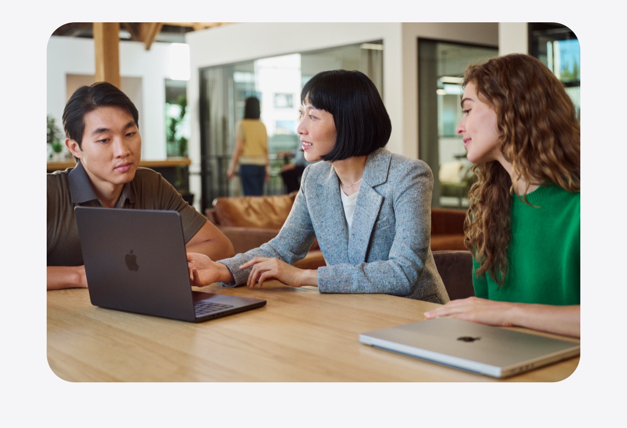 Three people collaborate around a MacBook at a table in an open workspace.