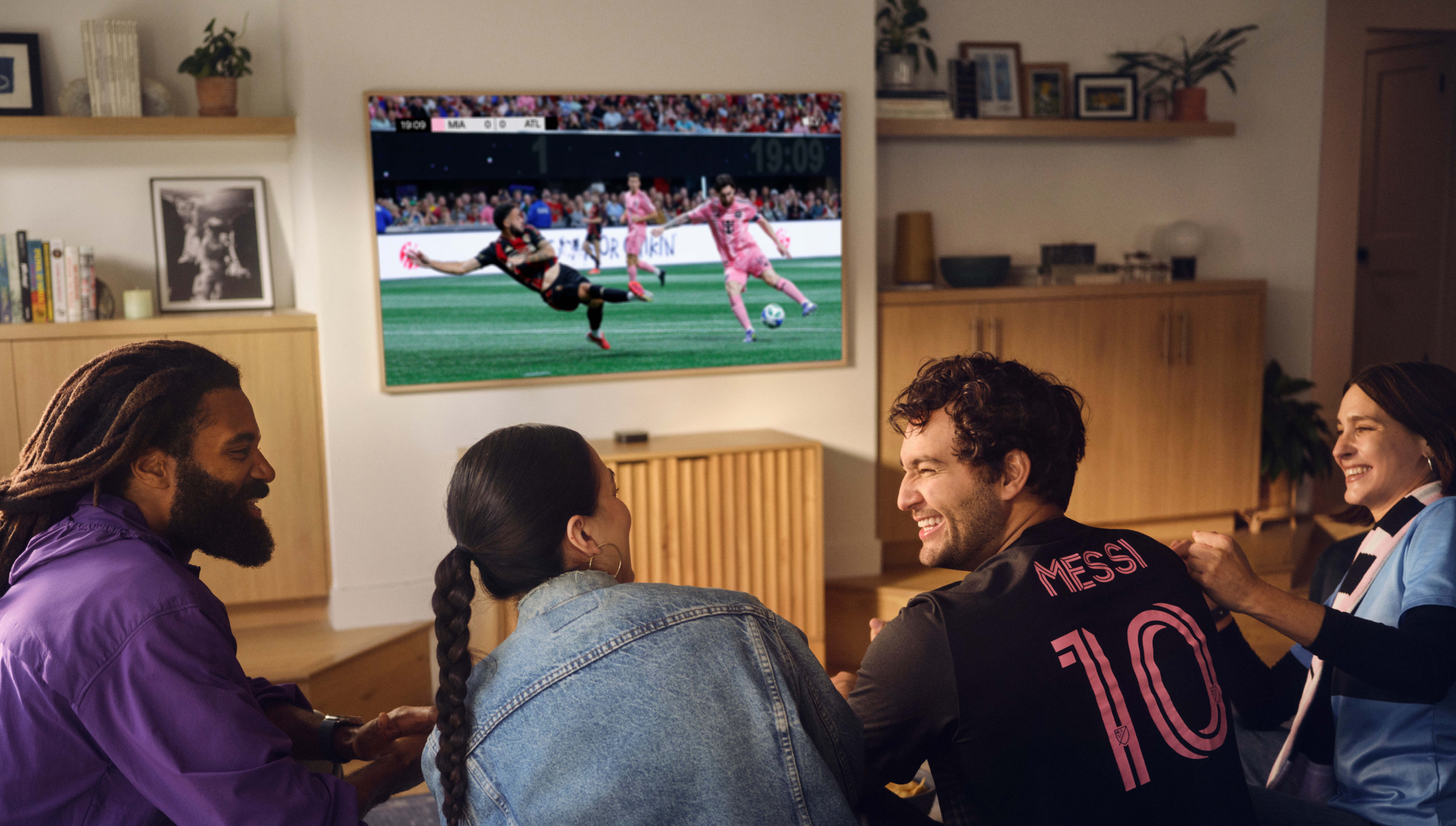 Группа из четырех улыбающихся друзей сидит на диване и смотрит MLS Season Pass по телевизору.