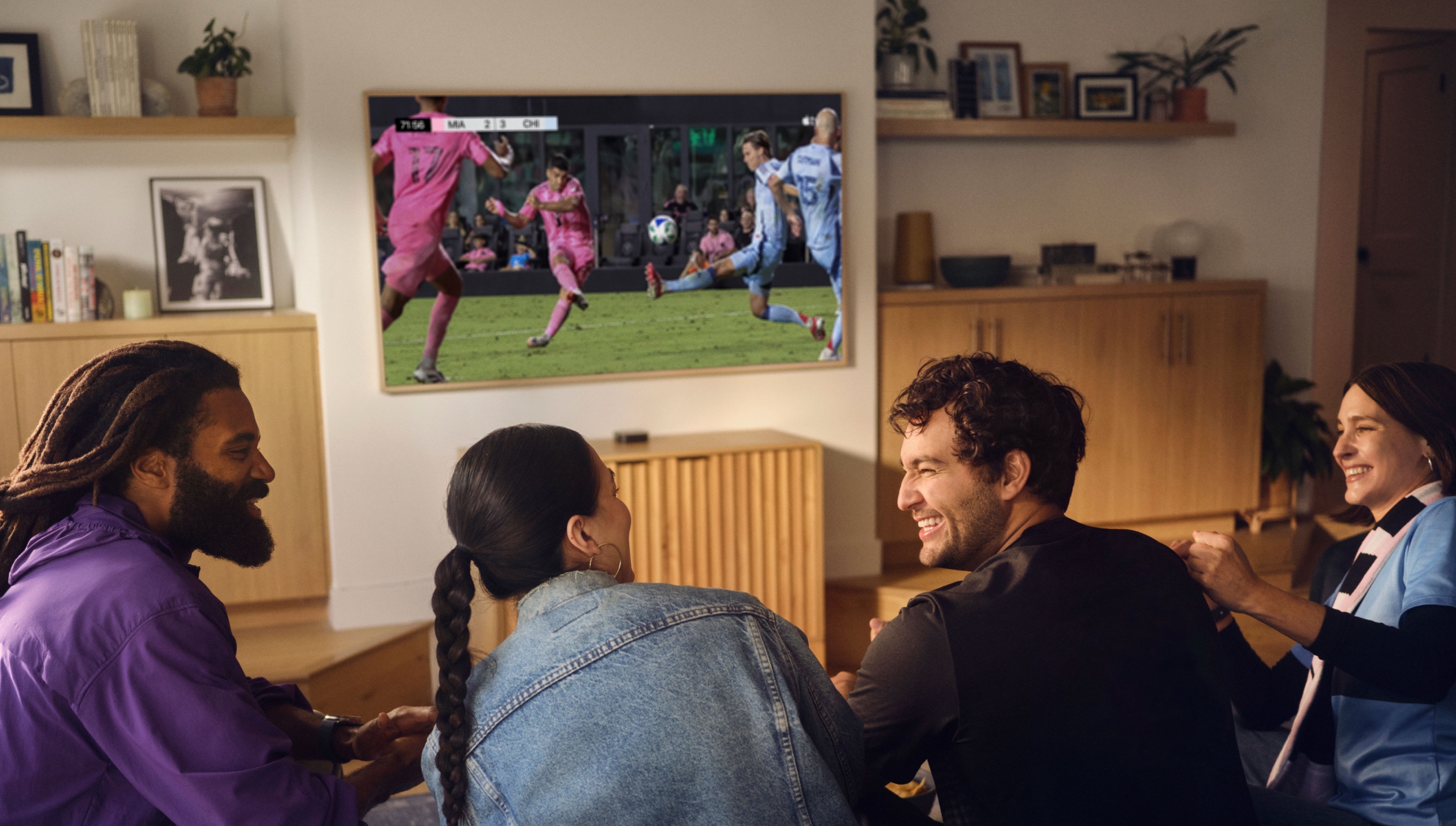 Un grupo sonriente de cuatro amigos sentados juntos en un sofá, viendo un partido de MLS en un televisor.