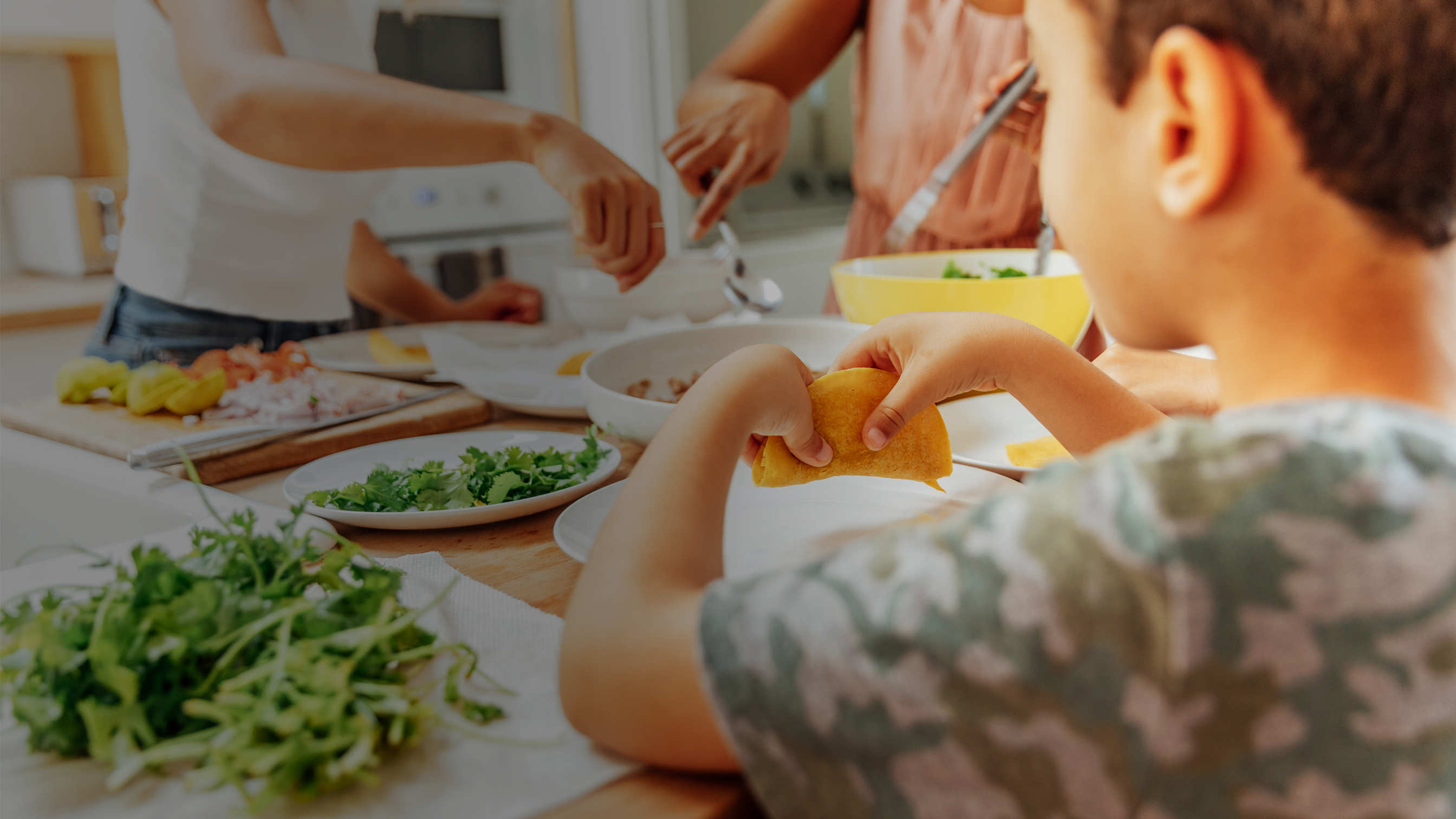 Uma criança desfrutando de uma refeição em uma mesa enquanto duas pessoas preparam comida.