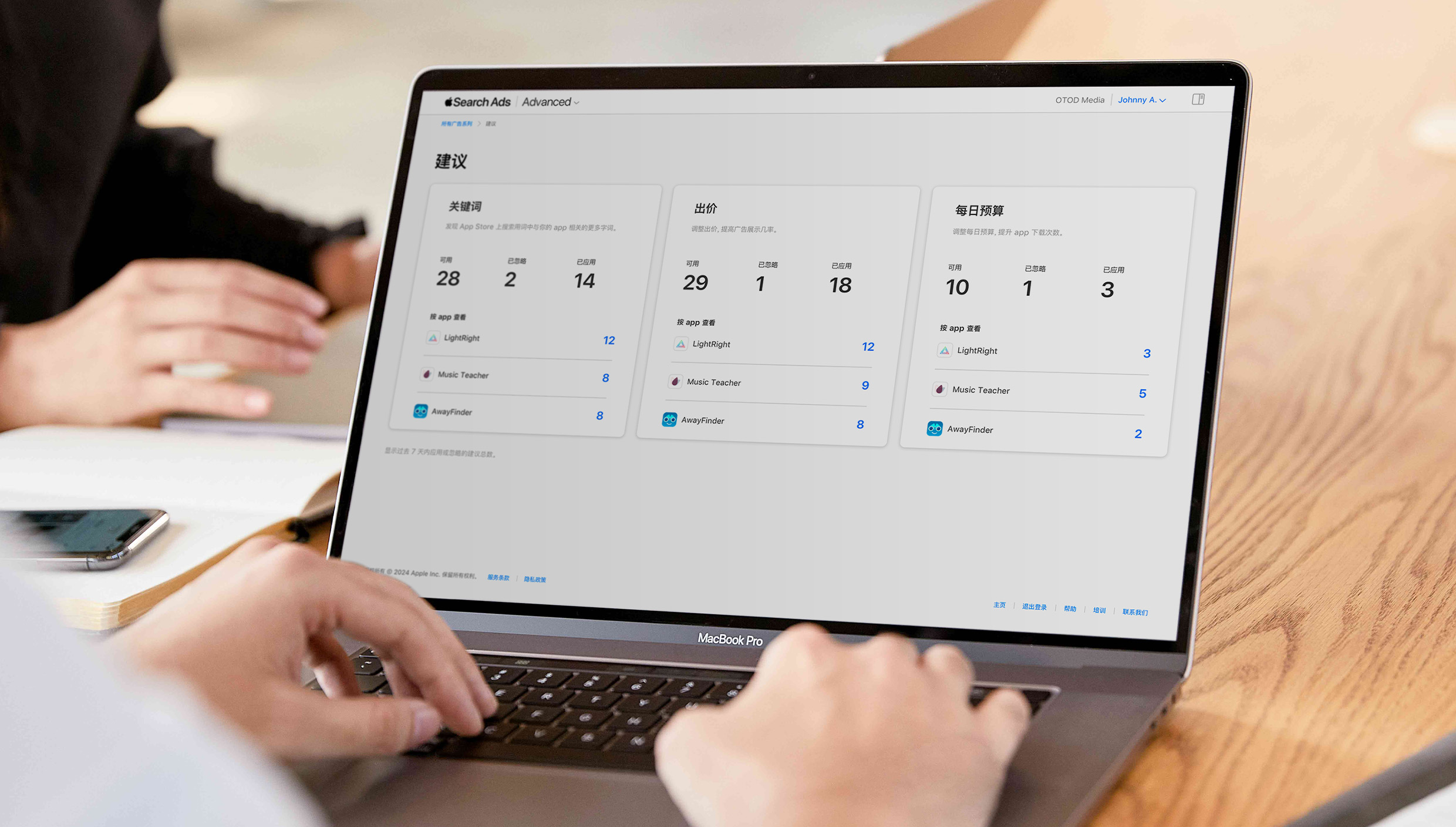 两只手在 MacBook 键盘上打字的特写。屏幕上打开了 Apple Search Ads“建议”页面，并显示按关键词、出价和每日预算划分的推荐。