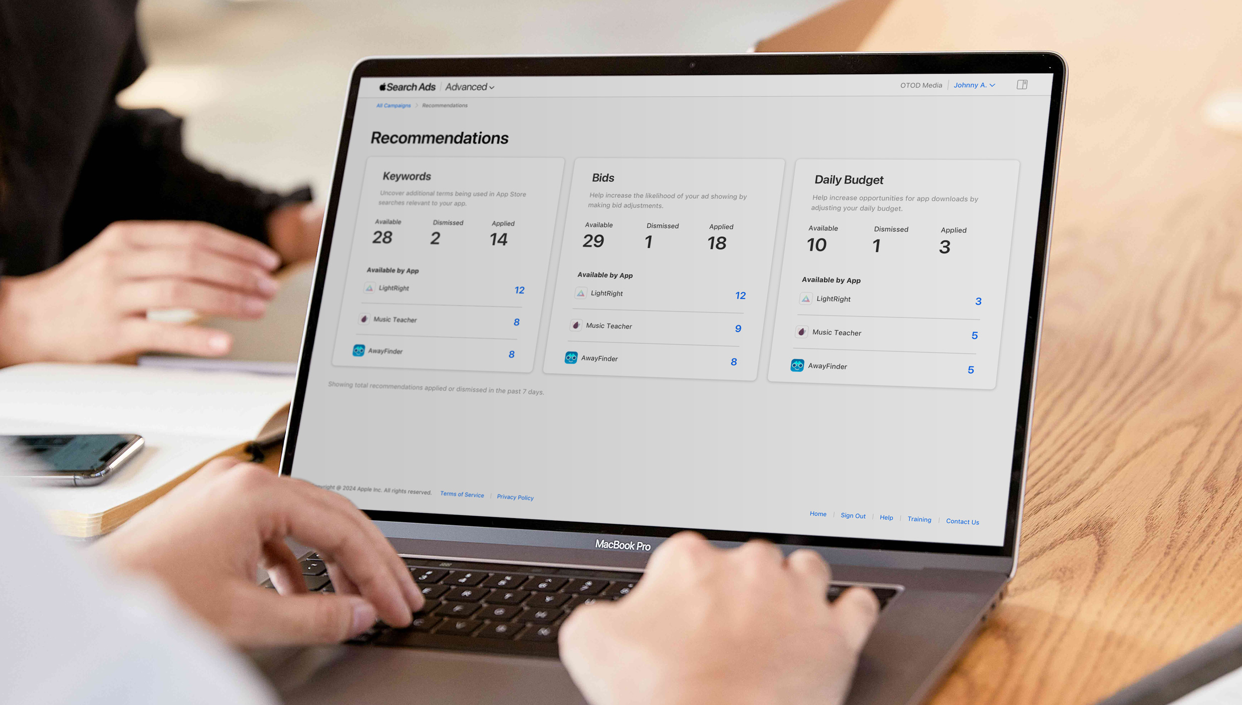 Close up of two hands typing on a MacBook keyboard. The screen is open to the Apple Search Ads Recommendations page and shows recommendations organized by keywords, bids, and daily budget.